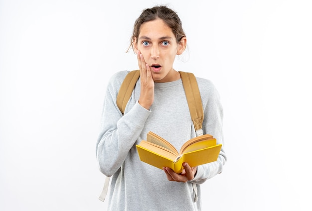 Estudiante joven asustado con mochila sosteniendo el libro poniendo la mano en la mejilla aislada en la pared blanca