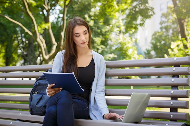 Estudiante joven en un banco. Chica morena haciendo la tarea en el parque con laptop y libro de texto. Concepto de estilo de vida, tecnología y educación.