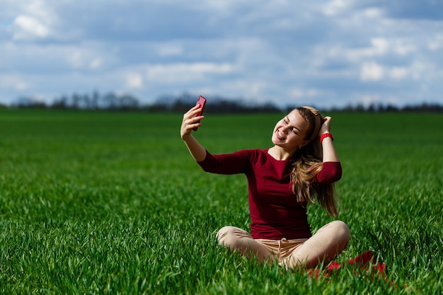 Estudiante joven y bella mujer con un teléfono en sus manos sentado en la hierba. Chica toma selfies y toma fotos selfie. Ella sonríe y disfruta de un día cálido. Foto de concepto en smartphone