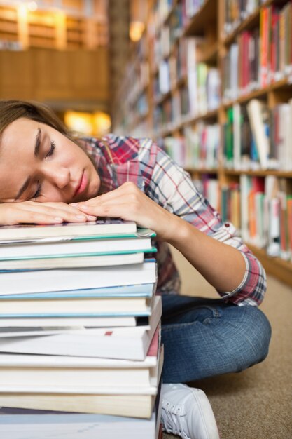 Estudiante joven dormitando que se sienta en el piso de la biblioteca que se inclina en la pila de libros