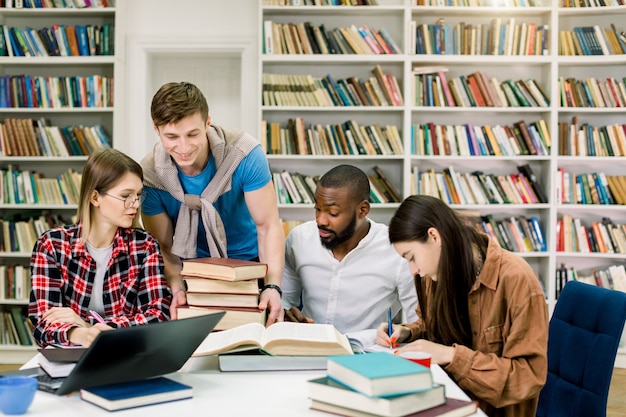 Estudiante joven guapo poniendo muchos libros diferentes sobre la mesa para sus amigos multirraciales de la universidad, sentados y estudiando juntos en la sala de lectura de la biblioteca del campus moderno.