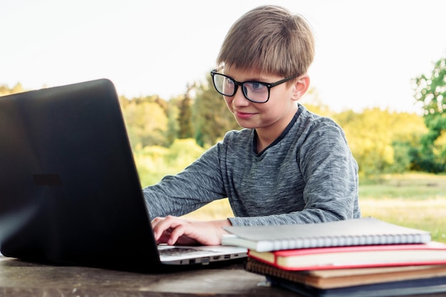 Estudiante joven inteligente con gafas cuadradas sonriendo mientras estudia con una computadora portátil en el parque Niño con manga larga gris mirando la pantalla del dispositivo con ojos atentos y disfrutando estudiando en línea