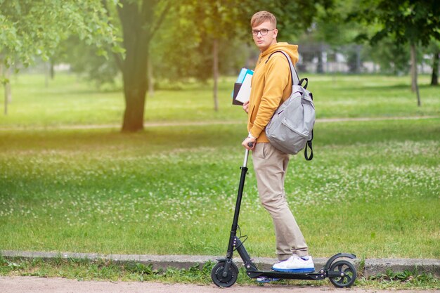 Estudiante joven con libros y mochila en scooter eléctrico en el parque