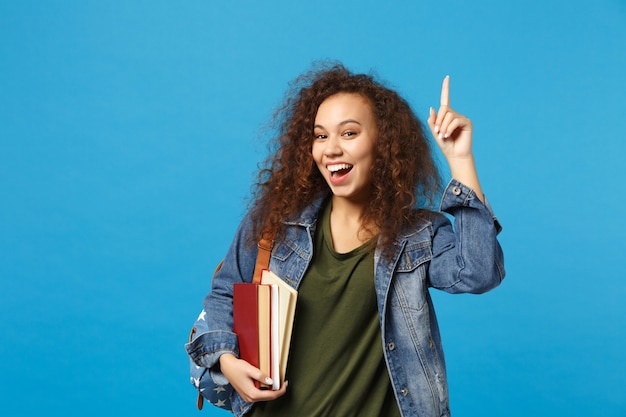 Estudiante joven en ropa de mezclilla y mochila tiene libros aislados en la pared azul