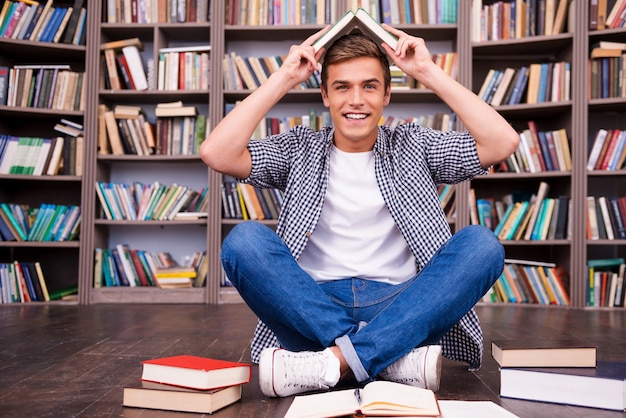 Estudiante juguetón. Juguetón joven llevando un libro en la cabeza y sonriendo mientras está sentado contra la estantería