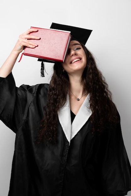 Estudiante con libro en toga de graduación y gorro listo para terminar la universidad. Mujer joven académica en vestido negro sonriendo.