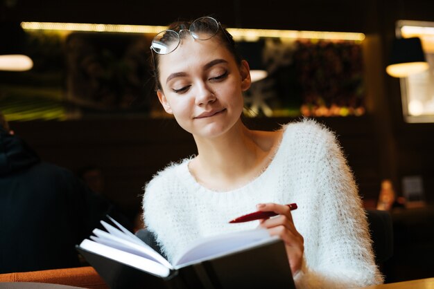 Foto estudiante linda joven en un suéter blanco se sienta en un café, escribe en su cuaderno