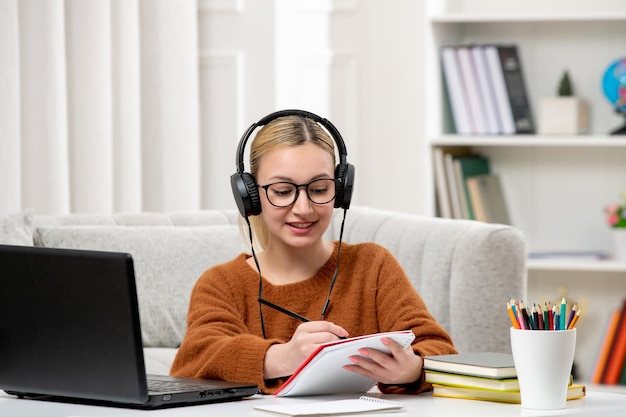 Estudiante en línea chica linda con gafas y suéter estudiando en la computadora escribiendo notas