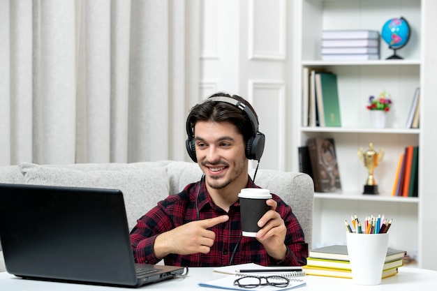 Estudiante en línea chico lindo en camisa a cuadros con gafas estudiando en la computadora sonriendo sosteniendo café