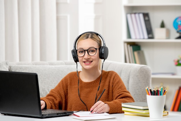 Estudiante en línea linda chica con gafas y suéter estudiando en la computadora sonriendo y escribiendo