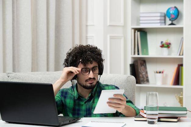 Estudiante en línea lindo joven estudiando en la computadora con gafas en camisa verde mirando notas