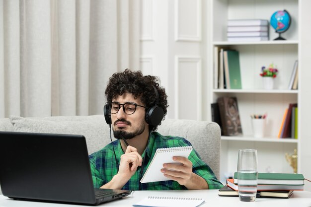 Estudiante en línea lindo joven estudiando en la computadora con gafas en camisa verde tomando notas