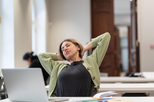 Estudiante madura cansada tomando un descanso estirando los brazos mientras estudia en línea en la biblioteca universitaria