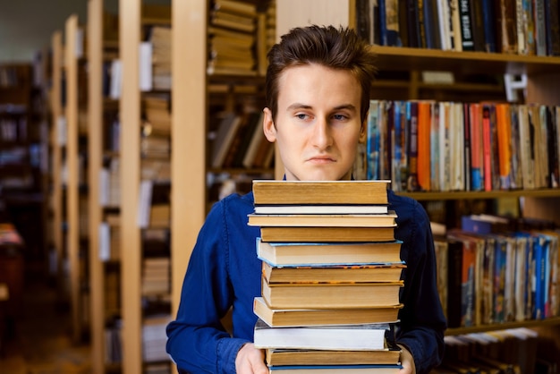 Estudiante masculino con imitación negativa y aburrida en su rostro sosteniendo una gran pila de libros en la biblioteca