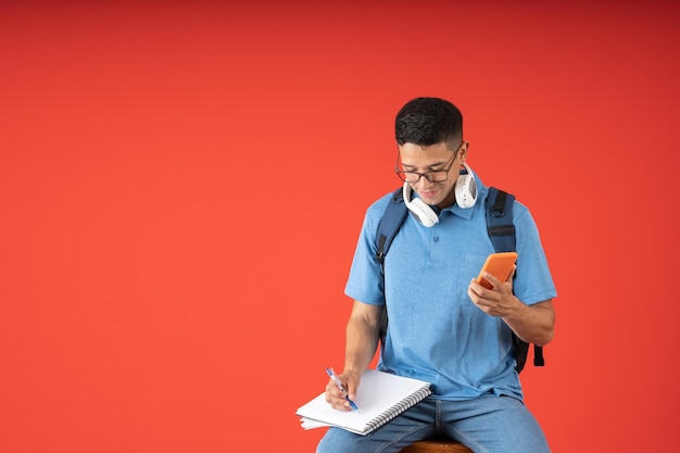 Estudiante masculino sentado escribiendo en su cuaderno con un bolígrafo mientras sostiene su teléfono celular