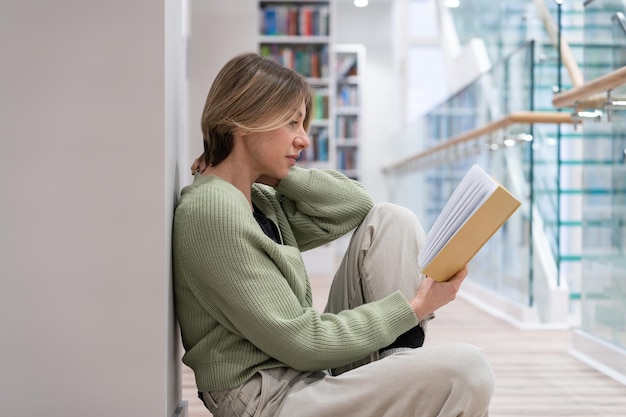 Estudiante de mediana edad sentado en el suelo leyendo un libro con una trama interesante en la biblioteca Educación