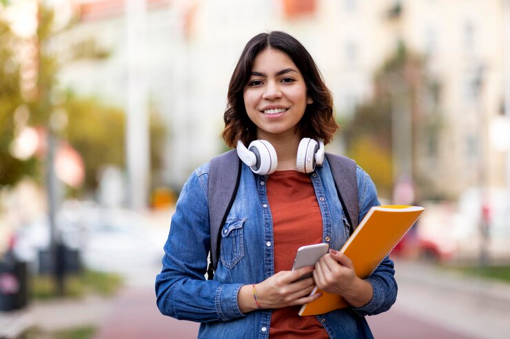 Estudiante moderno sonriente joven mujer árabe con libros de trabajo y teléfono inteligente posando al aire libre