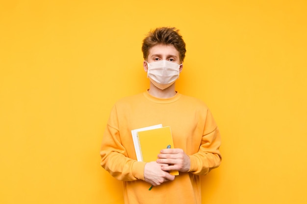 Estudiante molesto con una máscara médica sobre un fondo amarillo con libros y cuadernos en las manos