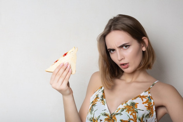 Estudiante morena enojada comiendo un sándwich sobre un fondo gris. Espacio para texto