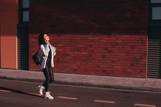 Estudiante morena de pelo rizado dando un paseo después de la escuela está hablando por teléfono y sosteniendo una computadora portátil