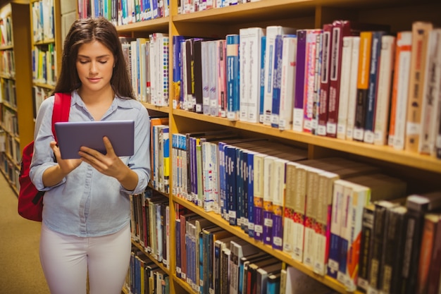 Estudiante morena que usa su tableta junto a los estantes de la biblioteca