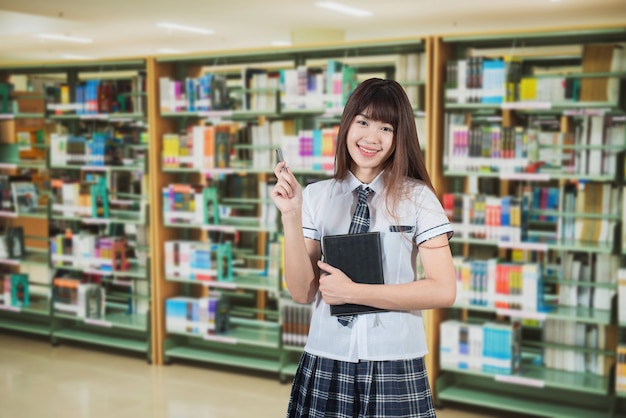 Estudiante de la muchacha asiática en el uniforme de Japón de la escuela en el fondo de la biblioteca