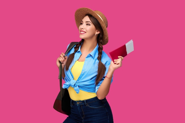 Estudiante mujer caucásica sonriente con pasaporte y boletos de avión. Chica en ropa casual y sombrero de paja. Viajero femenino sobre fondo rosa aislado.
