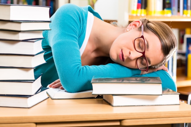 Foto estudiante - mujer joven en la biblioteca con libros de aprendizaje, ella está dormida, cansada y con exceso de trabajo