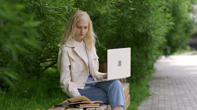 Estudiante mujer con laptop y libros se sienta en un banco del parque. Una mujer joven hojea libros y grabados en una computadora portátil sobre un fondo de árboles verdes. Días de estudio al aire libre. 4K UHD