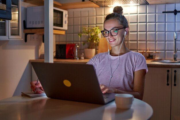 Estudiante mujer ocupada adicta al trabajo en auriculares preparándose para el examen, tomando notas en el cuaderno mientras ve el seminario web y aprendiendo una conferencia en línea en la computadora portátil a última hora de la noche