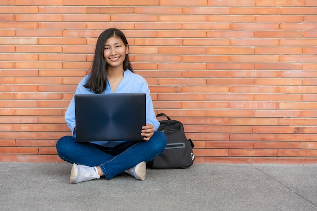 Estudiante de mujer sonriente asiático que sienta y que usa la computadora portátil en fondo del ladrillo en campus. Chica adolescente feliz estudiante de secundaria al aire libre.