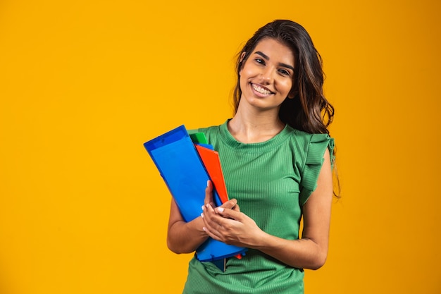 Estudiante mujer sonriente con libros escolares en manos sobre fondo amarillo.