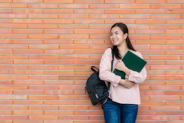 estudiante mujer sonriente sosteniendo libro posando sobre fondo de ladrillo en el campus.