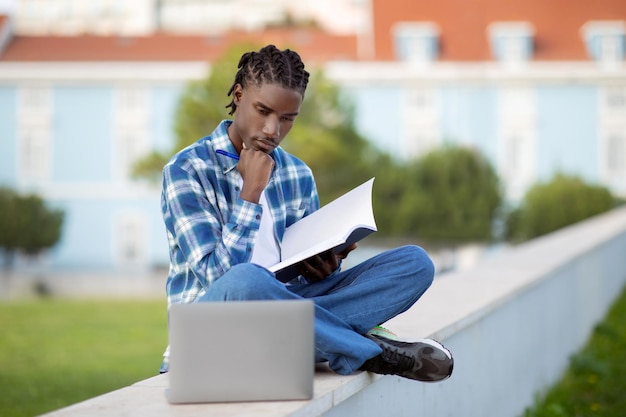 Estudiante negro aprendiendo con un libro usando una computadora portátil en el parque del campus