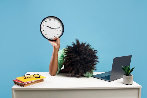 Estudiante negro cansado acostado en el escritorio con computadora portátil y libros sosteniendo el reloj y poniéndose la cabeza