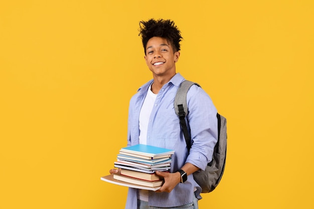 Estudiante negro feliz con mochila y una pila de libros sobre fondo amarillo