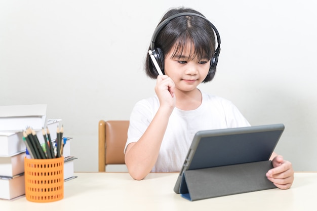 Estudiante de niña asiática alegre lleva auriculares escribe en tableta digital para estudiar en casa