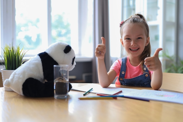 Estudiante de niña feliz estudia en línea en casa, niño pequeño sonriente que muestra el pulgar hacia arriba para recomendar clase o lección.