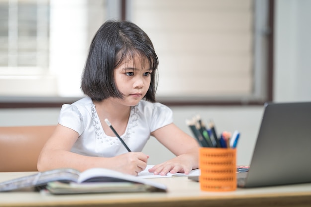 Estudiante de niña de niños asiáticos escribiendo en el cuaderno mientras estudia en línea a través de la computadora portátil mientras está en casa. Concepto de educación Foto de stock