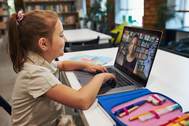 Estudiante niña pequeña teniendo clase de video remotamente con su maestro en la computadora portátil sentada en el escritorio