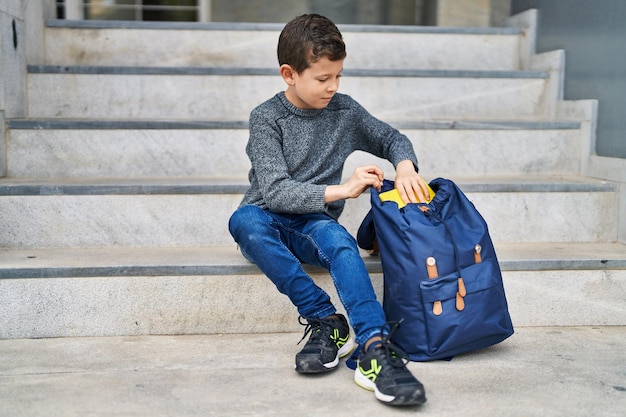 Estudiante de niño rubio sosteniendo un libro sentado en las escaleras en la escuela