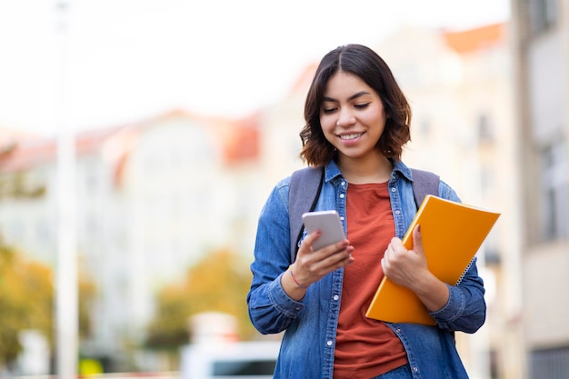 Estudiante de Oriente Medio caminando con teléfonos inteligentes y libros de trabajo en las calles de la ciudad