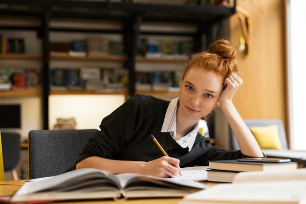 Estudiante pelirroja feliz sentado en la mesa con libros en la biblioteca escribiendo notas