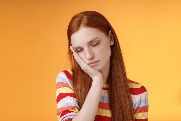 Estudiante pelirroja linda cansada agotada sentirse soñolienta quedarse dormida de pie inclinada cara palma cerrar ojos trabajando medio tiempo turno de noche, soñar despierto sin energía quiero dormir cama fondo naranja.