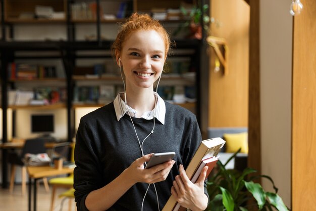 Estudiante pelirroja posando en el interior de la biblioteca sosteniendo libros, escuchando música con auriculares y usando el teléfono móvil