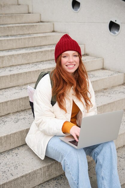 Una estudiante pelirroja sonriente se sienta en las escaleras al aire libre y usa una computadora portátil para conectarse a wifi público en la ciudad