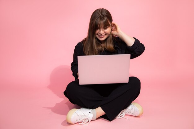 Estudiante de pelo castaño sorprendido encantadora chica en ropa casual sosteniendo ordenador portátil sentado sobre fondo rosa. Concepto de educación en línea. Foto de estudio. Mirando la computadora portátil.