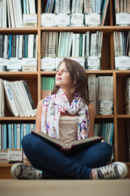 Estudiante pensativa contra la estantería con un libro en el piso de la biblioteca