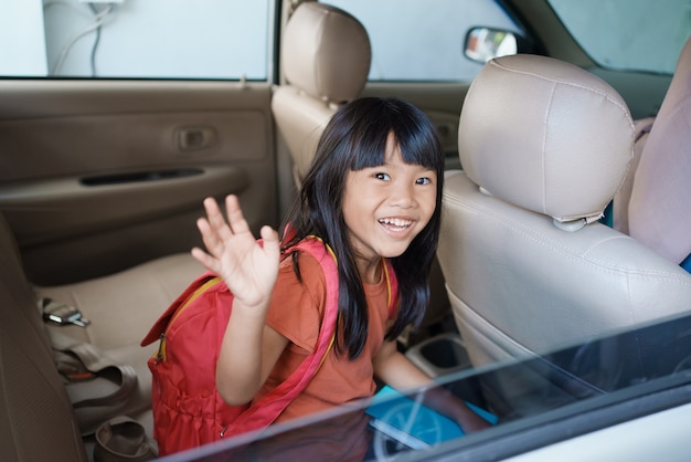 Estudiante de primaria asiática sentado en el coche y diciendo adiós mientras va a la escuela por la mañana