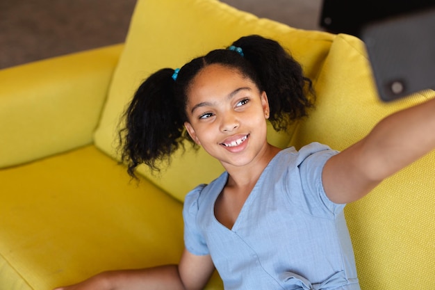 Una estudiante primaria biracial sonriente tomando una selfie mientras está sentada en el sofá de la escuela